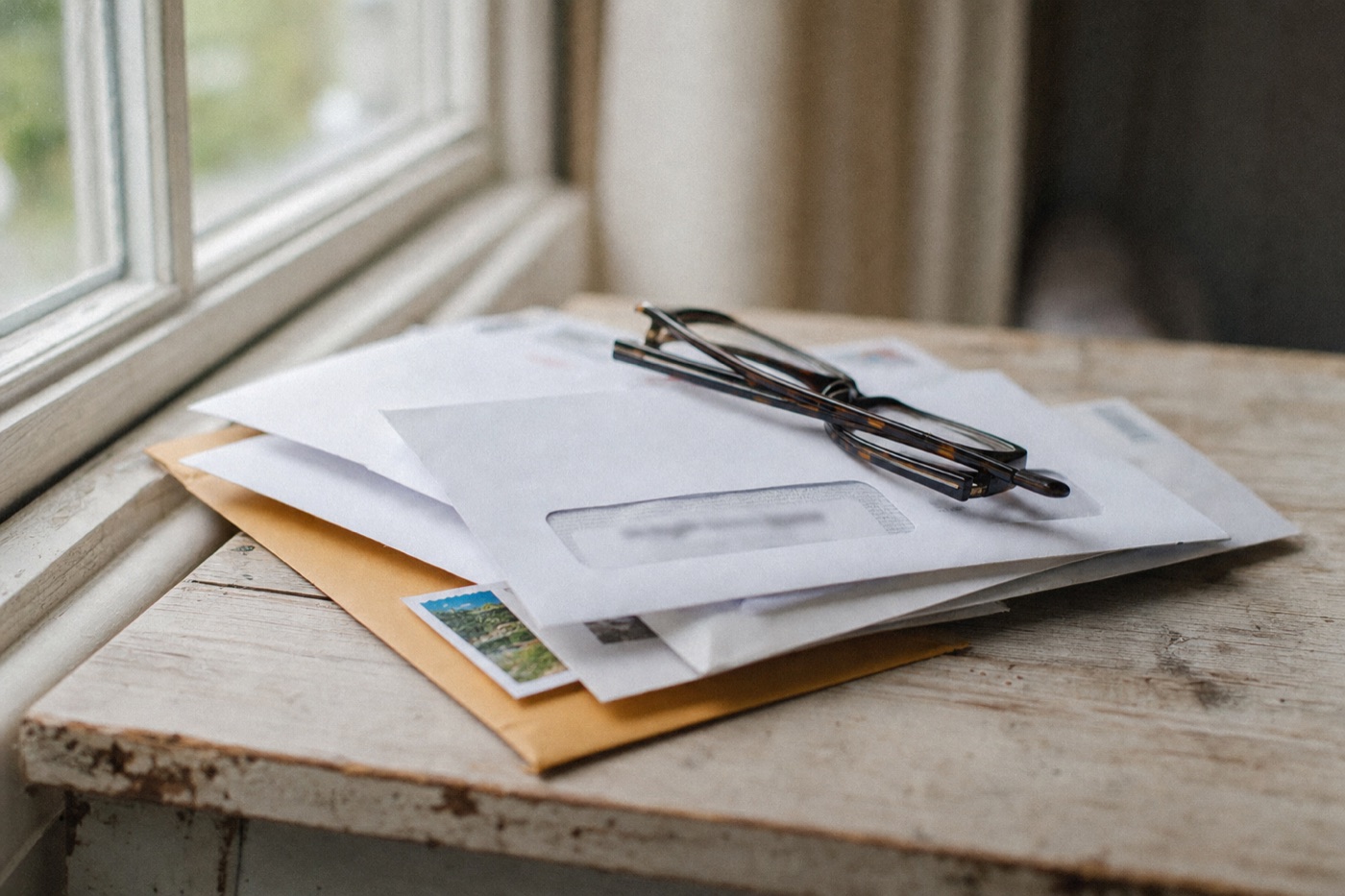 Generated image: a stack of unopened mail with folded reading glasses on top, on a worn table by a window.